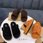 A display of black, brown, and tan suede slides with white stitching on a round table, with boxes and a Zara bag in the background.