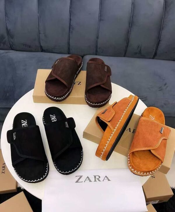 IMG-20260708-WA0045 A display of black, brown, and tan suede slides with white stitching on a round table, with boxes and a Zara bag in the background.