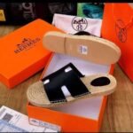 A pair of black leather Hermes Paris sandals with jute sole in an open orange box, with a dust bag and box in the background, showcasing luxury footwear.