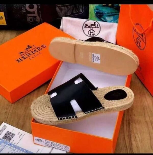 A pair of black leather Hermes Paris sandals with jute sole in an open orange box, with a dust bag and box in the background, showcasing luxury footwear.