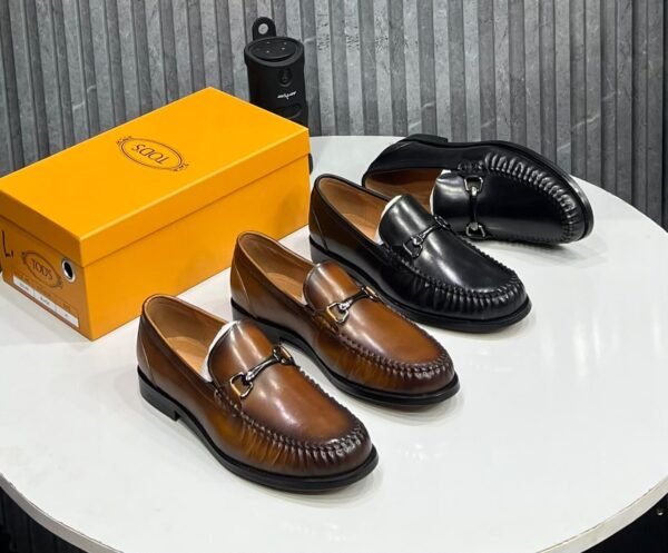 A stylish display of brown and black leather loafers with buckle details, accompanied by a yellow shoe box on a white table.