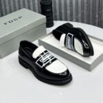 A pair of black and white loafers with bold design, displayed on a white table with a gray shoe box and a black cylindrical object in the background.