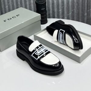 A pair of black and white loafers with bold design, displayed on a white table with a gray shoe box and a black cylindrical object in the background.