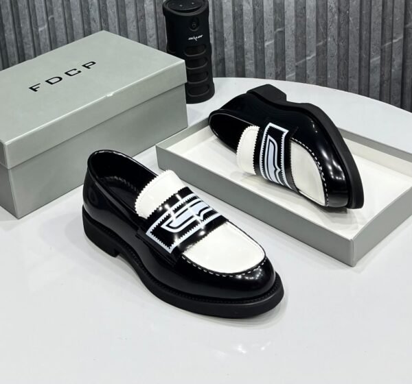 IMG-20260708-WA0048 A pair of black and white loafers with bold design, displayed on a white table with a gray shoe box and a black cylindrical object in the background.
