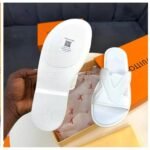 A close-up shot of a pair of white slide sandals with a Louis Vuitton-inspired design, showcasing the sole and the shoe's sleek design, with a hand holding the sandal and a box in the background.