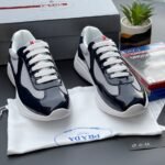 A pair of navy blue and white Prada sneakers on a white cloth bag with a Prada logo, surrounded by a shoe box, a plant, and a PS4 game.