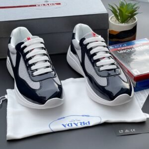 A pair of navy blue and white Prada sneakers on a white cloth bag with a Prada logo, surrounded by a shoe box, a plant, and a PS4 game.