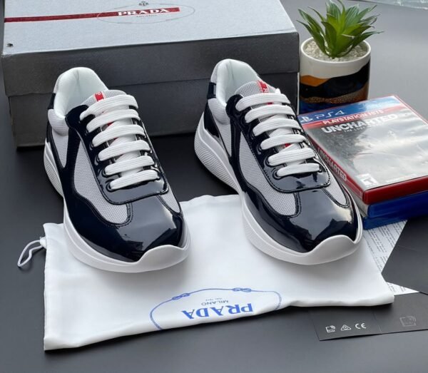 A pair of navy blue and white Prada sneakers on a white cloth bag with a Prada logo, surrounded by a shoe box, a plant, and a PS4 game.