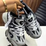 A pair of grey and black Adidas sneakers with white laces and tags, held by a hand with a gold bracelet, showcasing the shoe's details and design.