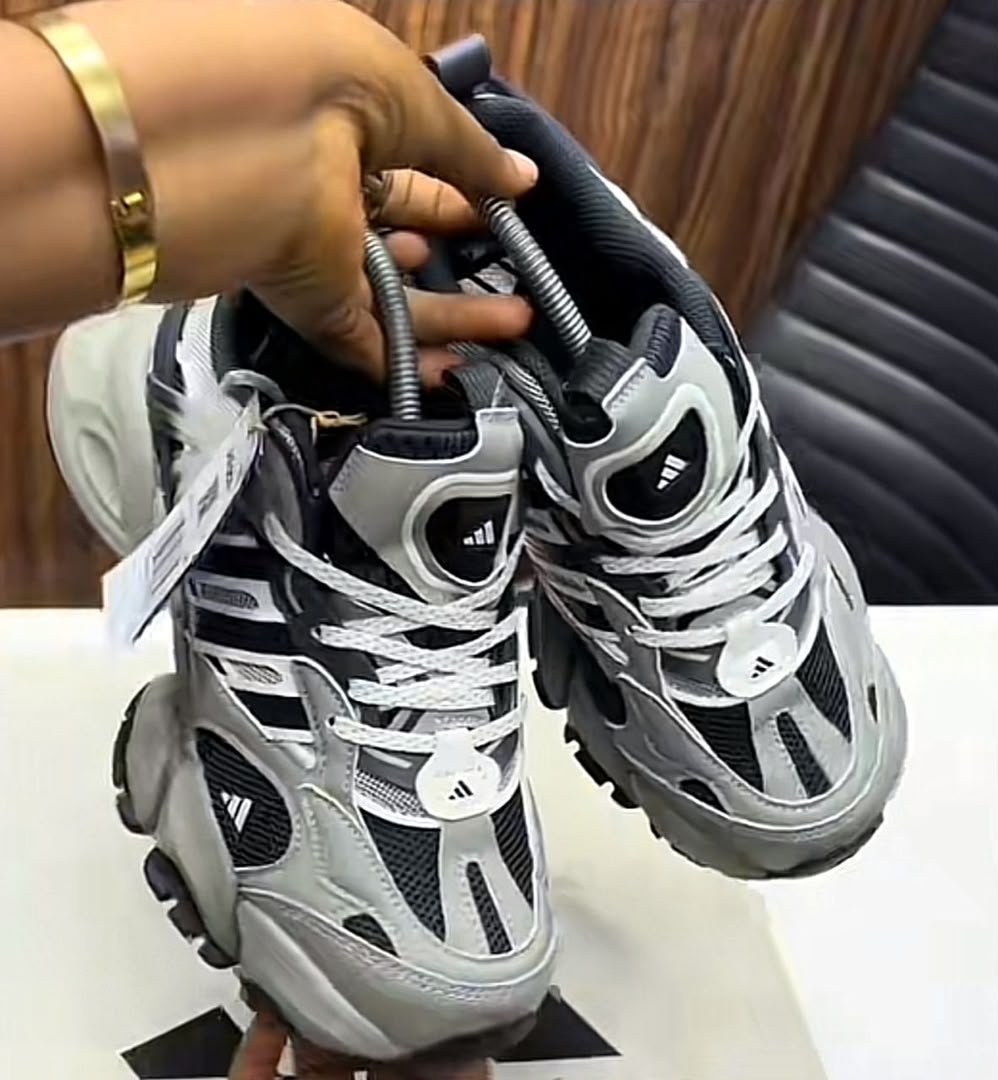 A pair of grey and black Adidas sneakers with white laces and tags, held by a hand with a gold bracelet, showcasing the shoe's details and design.