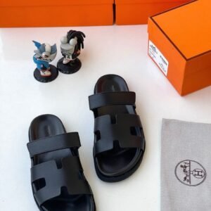 Luxurious black slides with two wide straps, presented with orange shoe boxes and accessories, embodying JC Luxury Footwears' high-end style.