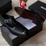 High-quality black leather monk strap shoes with a sleek buckle and ventilation holes, presented on a black shoe box with a reflective surface.