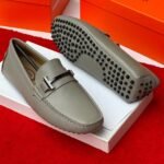 A pair of stylish grey leather loafers with black studs on the sole, displayed on a white shoe box with an orange background, showcasing Jsfootis luxury footwear.