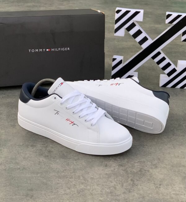 root A pair of white Tommy Hilfiger sneakers with black and red logo, displayed with a black box on a concrete floor.