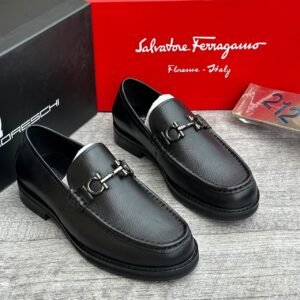 A pair of sleek black leather loafers with metal buckles, displayed on a wooden surface with Salvatore Ferragamo and Jsfootis branding in the background, showcasing luxury footwear.