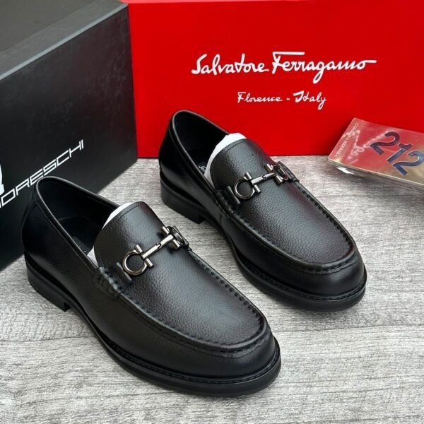 A pair of sleek black leather loafers with metal buckles, displayed on a wooden surface with Salvatore Ferragamo and Jsfootis branding in the background, showcasing luxury footwear.