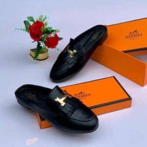 A pair of black leather loafers with gold-toned buckles, displayed on top of and next to orange shoe boxes with the Hermes Paris logo, accompanied by a small vase of red roses.