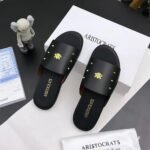 A pair of black slides with gold accents, placed on a white box and paper with the brand name 'Aristocrats' and a small figurine nearby.