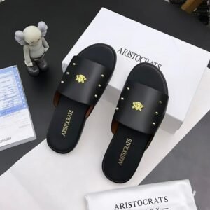 A pair of black slides with gold accents, placed on a white box and paper with the brand name 'Aristocrats' and a small figurine nearby.
