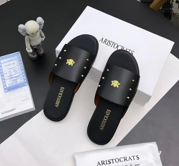 root A pair of black slides with gold accents, placed on a white box and paper with the brand name 'Aristocrats' and a small figurine nearby.