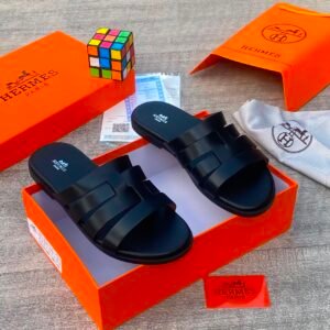 A pair of black Hermes slides nestled in an orange box, surrounded by luxury accessories.