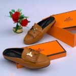A pair of black leather loafers with gold-toned buckles, displayed on top of and next to orange shoe boxes with the Hermes Paris logo, accompanied by a small vase of red roses.