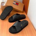A pair of black slides with a gold logo on the strap, displayed on a wooden table with a shoe box in the background.