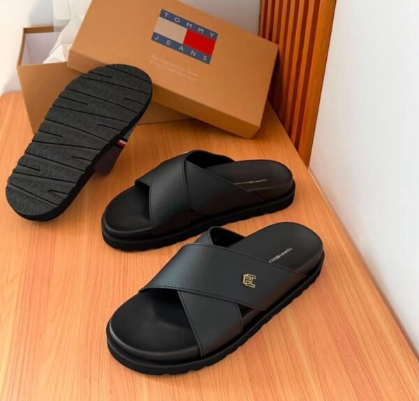 root A pair of black slides with a gold logo on the strap, displayed on a wooden table with a shoe box in the background.