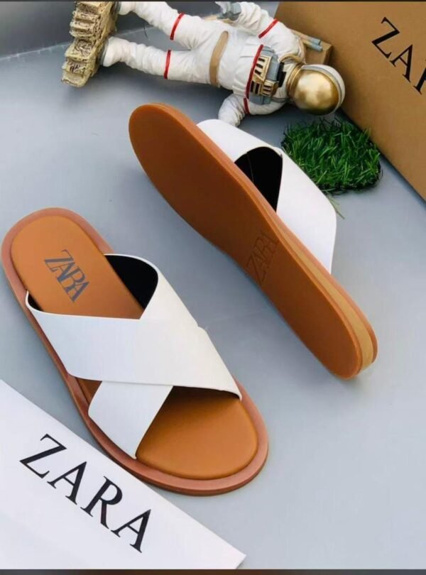root A pair of white ZARA slides with brown soles, displayed on a gray surface with a small astronaut figurine and a ZARA box in the background.