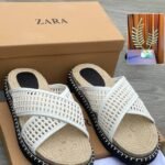A pair of white slides with woven straps and black outsole, placed on a brown shoe box and cloth with the ZARA logo.