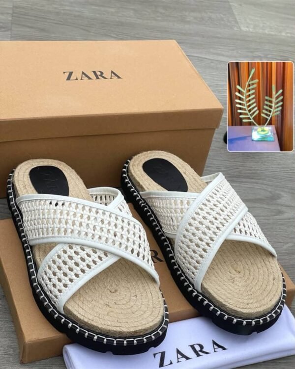 root A pair of white slides with woven straps and black outsole, placed on a brown shoe box and cloth with the ZARA logo.
